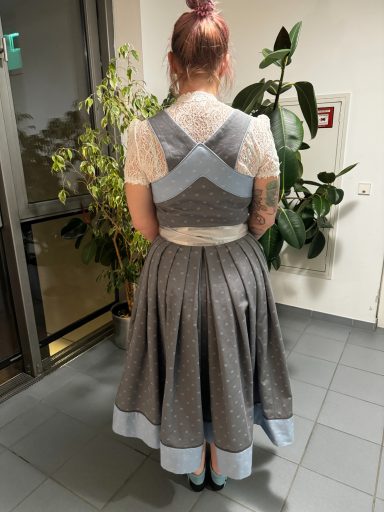 Frau mit geflochtenem Haar trägt ein traditionelles, graues Kleid mit blauen Akzenten.